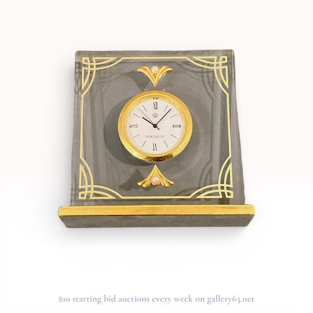 Description
Mikimoto International quartz desk clock, late 20th century. The clear rectangular acrylic case features gold-tone etched scrollwork at the corners and two fan-shaped mounts each set with a single cultured pearl. A white circular dial is housed in a gold-tone bezel with Roman numerals at the cardinal hours and is mounted to a rectangular gold-tone metal base. The dial and the reverse of the movement housing are both marked Mikimoto International.