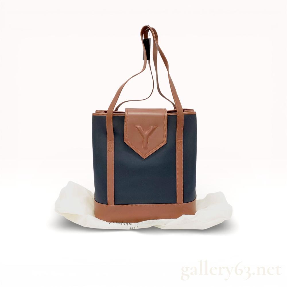 Description
Yves Saint Laurent tote bag having a structured nylon body with leather trim and base. Design featuring leather shoulder straps, leather corner reinforcements, and a leather front flap accent embossed with YSL motif. Open-top tote format with interior textile lining and branded tag. Bag presenting a contrasting two-tone construction combining nylon panels with smooth leather detailing throughout.