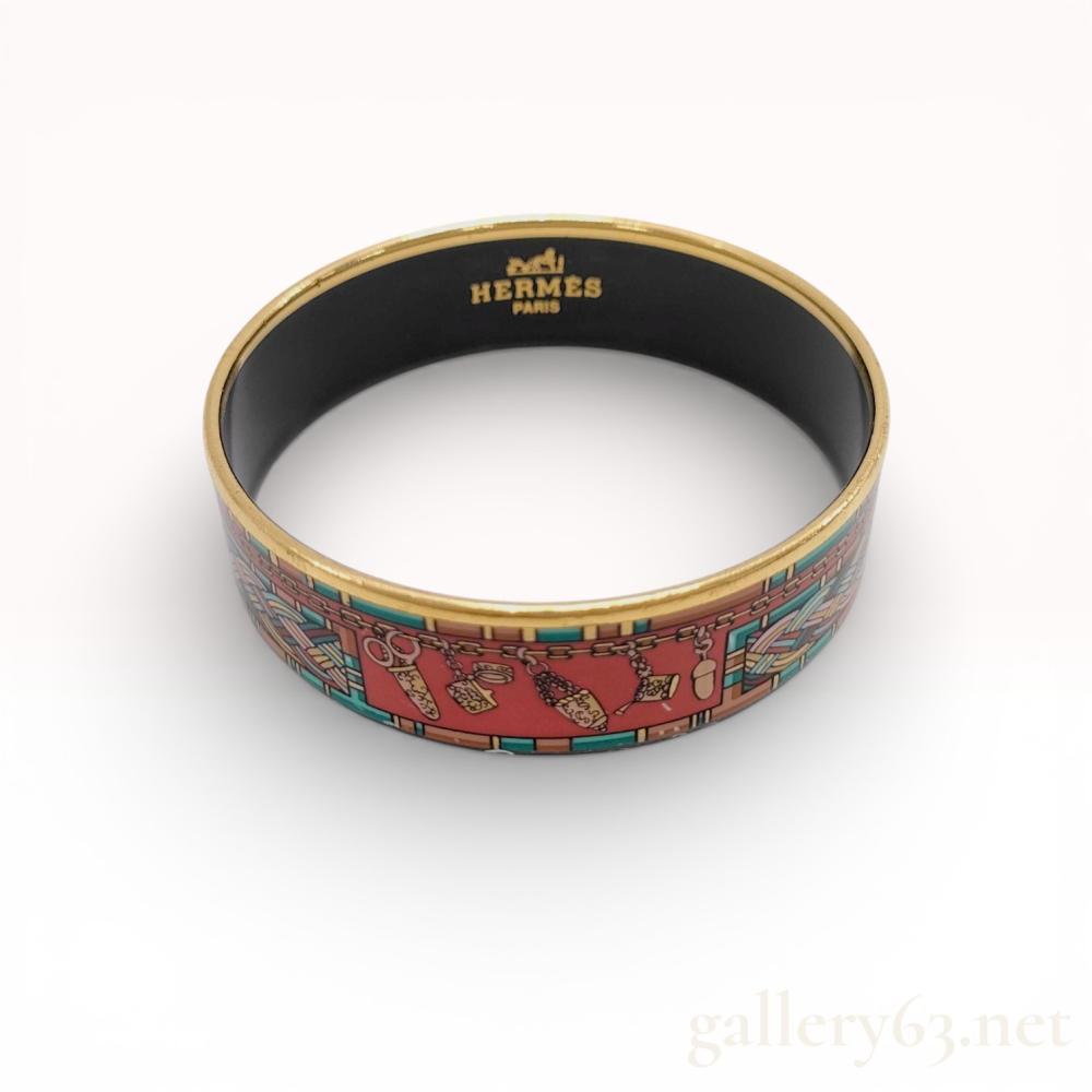 Description
Hermes wide enamel bangle featuring a printed design of sewing implements including scissors and thimbles on a red ground. The central motif is framed by a multicolored braided border and set in a gold-toned metal mount. The interior is finished with a black coating and bears the gilded Hermes Paris logo with horse and carriage, along with a Made in Austria mark.
Width: 20 mm. Size: 19.