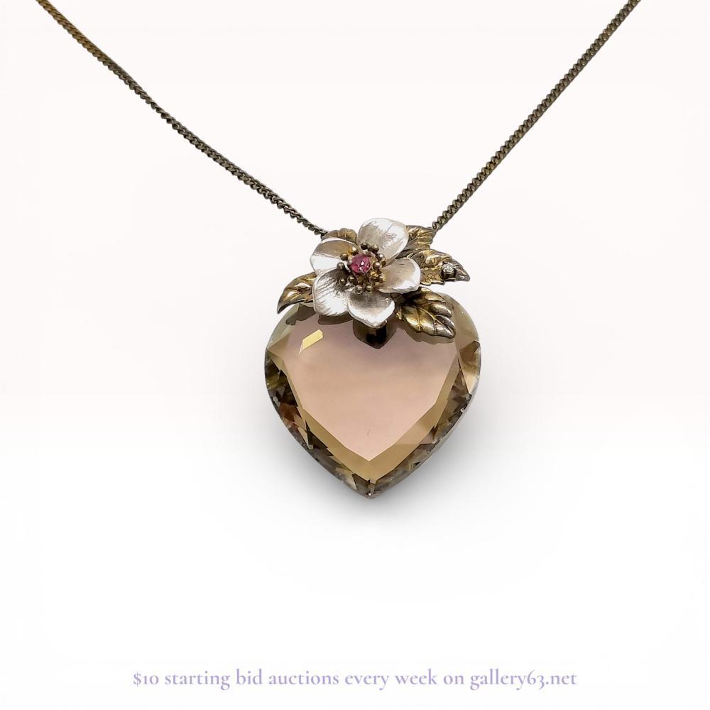 Description
Sterling silver perfume vial necklace featuring a heart-shaped faceted smoky-brown stone reservoir, likely smoky quartz. The vial is topped with a decorative enameled floral stopper accented by a pink center stone and gold-plated leaves set with colorless crystals. It is suspended from a fine curb-link chain with a spring ring clasp. The clasp tag is stamped SILVER 925.
Pendant: 35mm x 26mm
Chain length: 15.5 inches.