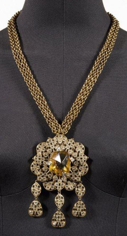 YVES SAINT LAURENT

Somptueux pendentif d’influence indienne en métal doré serti d’une importante pierre en résine façon citrine au centre entourée de boteths sertis de strass à l’imitation brillants, rehaussés de pendeloques à l’identique, monté sur une double chaîne torsadé. Signé