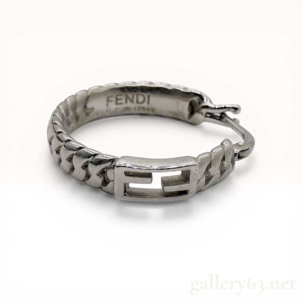 Description
Fendi single hoop earring from the Baguette collection, contemporary. The silver-tone metal hoop features a textured curb-link design with a central rectangular plaque displaying an open-work FF Zucca logo. It is secured with a hinged snap post closure and is stamped FENDI, MADE IN ITALY, and 1A4021.
Diameter: 25mm.