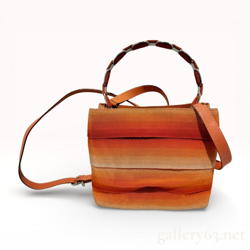 Description
Salvatore Ferragamo bucket bag featuring a tiered exterior of horizontal ombre-dyed grosgrain fabric strips transitioning from deep orange to cream. The structure is defined by an orange leather base and trim, featuring an articulated silver-tone metal and fabric top handle and a detachable leather crossbody strap. The interior is lined in black Gancini monogram jacquard fabric with a single zippered pocket and a silver-tone post-and-snap closure. An interior leather tag is silver-stamped with the Salvatore Ferragamo script, serial number AU-21 7184, and Made in Italy.
7.5 inches W x 7 inches H x 3.5 inches D.