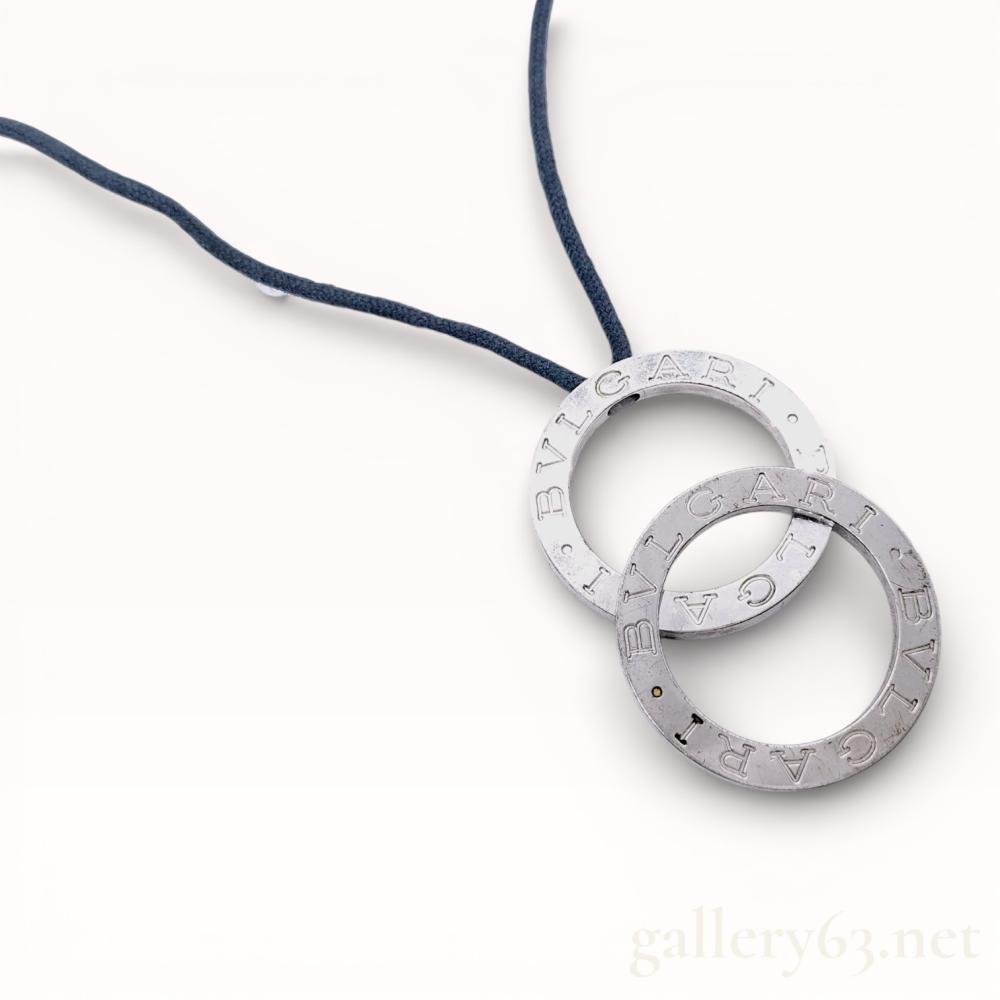 Description
Bvlgari sterling silver pendant necklace from the Bvlgari Bvlgari collection. The design features two interlocking flat circular pendants, known as the Silver in Silver model, suspended from a dark navy braided textile cord finished with silver-tone metal aglets. Each ring is engraved with the BVLGARI logo repeating twice on both faces. Includes an unbranded black velvet drawstring pouch.