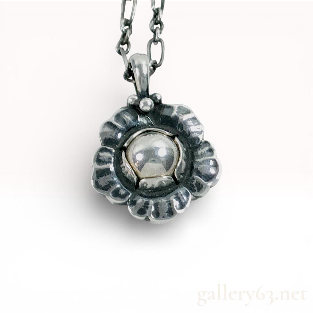 Description
 georg jensen necklace, authentic. sterling silver pendant shaped like a flower with rounded central dome and scalloped petal border. back stamped â2002â and â925 S georg jensen denmark.â attached to medium-length silver link chain with spring ring clasp. pendant and chain both show light tarnish and minor surface wear. dimensional construction gives the floral form depth. polished highlights contrast with oxidized recesses, emphasizing the petal detailing.