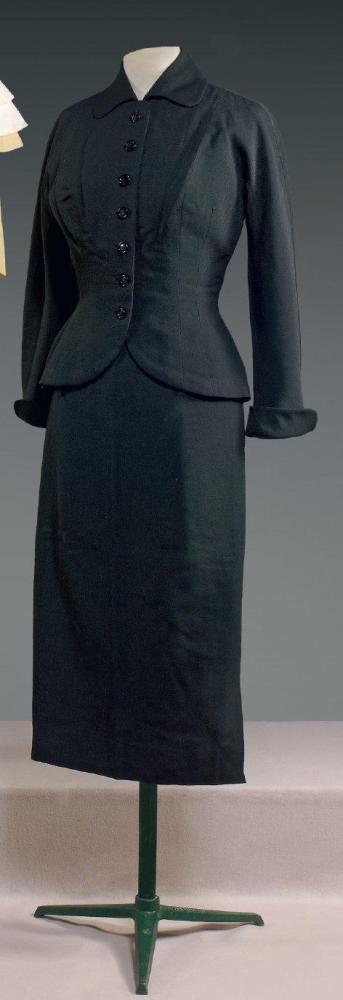 Emilio SCHUBERT Roma, circa 1950

Tailleur en toile de laine noire, veste courte cintrée, col claudine, simple boutonnage,parementures finissant par un arrondi, manches longues à petit revers rappelant le col, sur les petites hanches au dos effet de deux poches passepoilées en biais, jupe droite à longueur mi mollet, petite fente plate sur un côté. Griffe noire, graphisme blanc