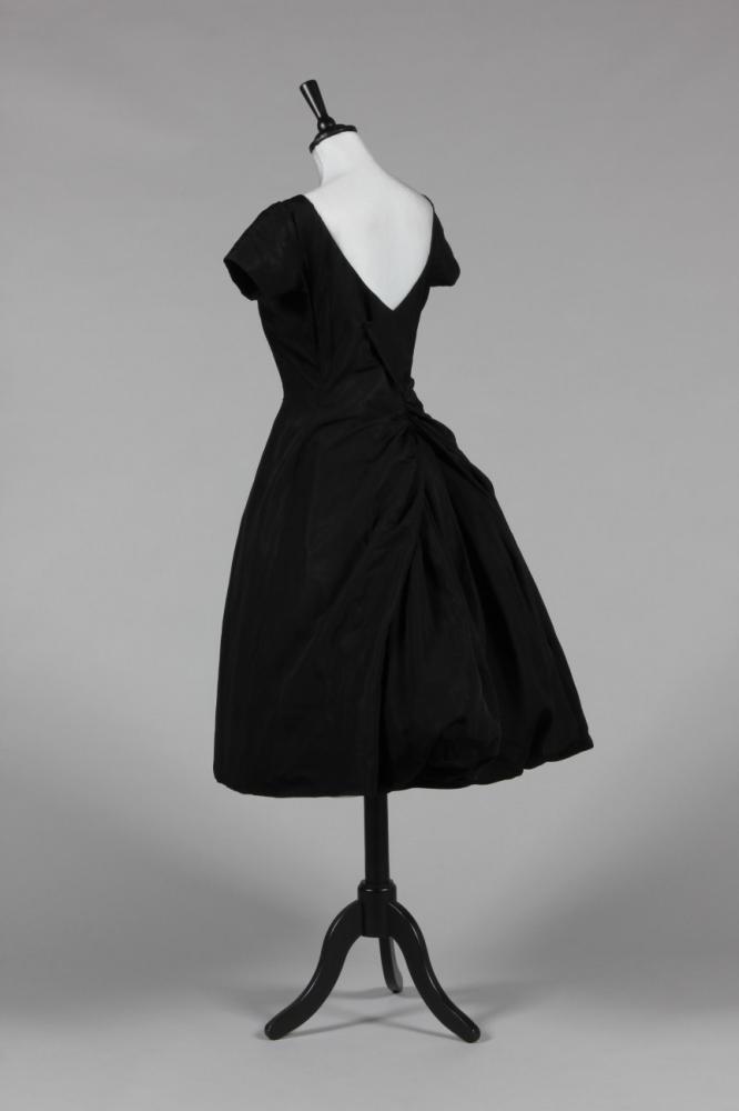 

An Yves Saint Laurent for Christian Dior couture black faille cocktail dress, probably Spring-Summer, 1958, un-labelled, with princess-line bodice and skirt, the skirt sweeping round to the back waist which frame vertically draped flounces centre-back, creating a bustle-like effect, with cross-over closure panel to back bodice, integral tulle corset with suspenders, layered tulle petticoats, bust approx 86cm, 34in, waist 66cm, 26in
