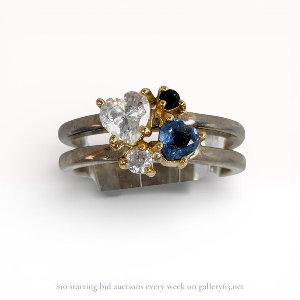 Description
Contemporary multi-stone cluster ring featuring a two-tone construction of 18 karat gold and sterling silver. The design includes a heart-shaped clear stone, a heart-shaped blue stone, and two round accent stones in clear and dark blue, all prong-set in yellow gold atop a silver split-shank band. The interior of the shank is stamped with K18 and 925 hallmarks alongside a stylized maker’s mark.
Interior diameter: approximately 16.5mm
Ring size: US 6.