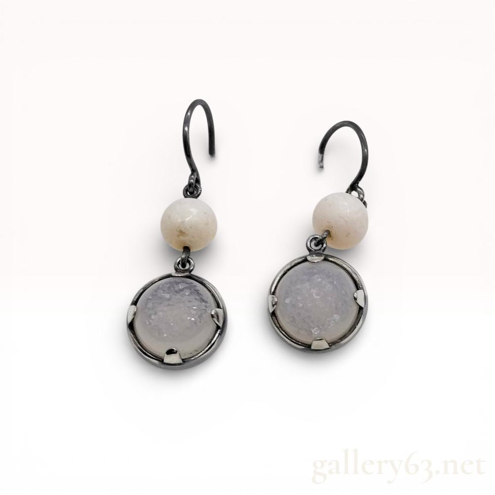 Description
A pair of matched drop earrings in darkened sterling silver. Each earring features a two-tiered dangle design, starting with a spherical off-white stone bead suspended from a shepherd’s hook ear wire. The lower drop consists of a circular pale lavender druzy quartz cabochon. The quartz is set within a bezel frame accented by four decorative prongs. The metal features an intentional aged patina. No hallmarks are visible. Total drop length is approximately 40mm. Weight: Not provided.