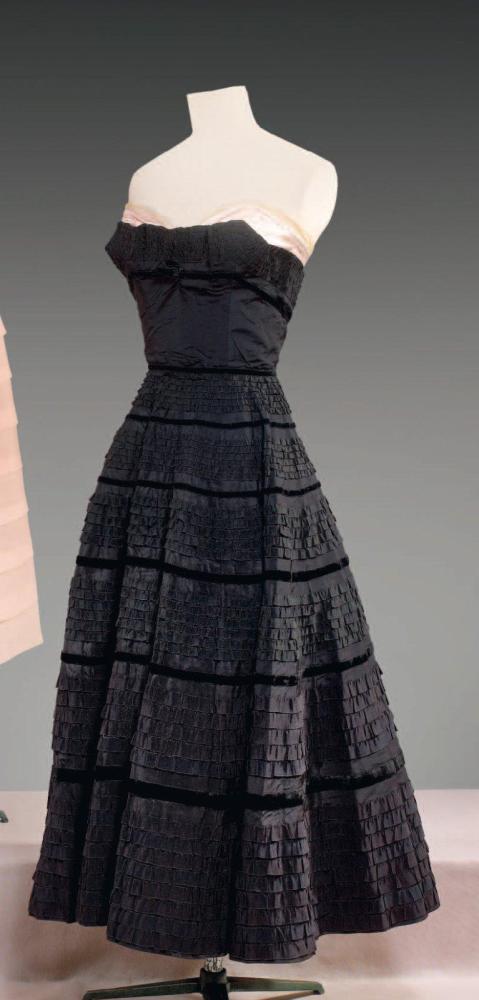 Emilio SCHUBERT Roma, circa 1950

Robe de cocktail en reps noir, haut bustier orné de gros grain bouillonés soulignés d’un ruban de velours, effet de corset en satin rose dépassant du bustier ,jupe très ample ornée d’une multitude de volants également de gros grain posés à la main avec rappel des rubans de velours, travail décroissant. Sans griffe