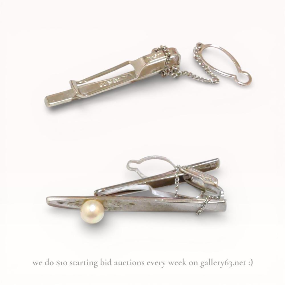 Description
Tie pin pair crafted in sterling silver, each featuring a linear bar form with polished surfaces. One pin set with a single round white pearl accent, the other presented in a plain streamlined design. Both pieces fitted with chain and loop backs for secure attachment. Presented together as a matched accessory group with a combined total weight of approximately 11.2 grams.