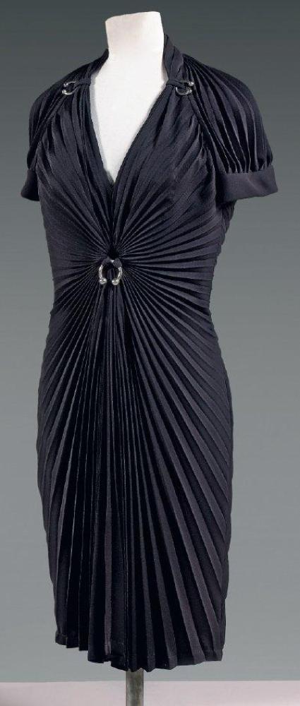 Thierry MUGLER, circa 1985

Robe de cocktail en crêpe noir à plissé soleil, décolleté en pointe agémenté de chaque côté d’anneaux ouverts, manches courtes raglan, rappel de l’anneau au niveau de la taille, jupe souple. Griffe bleue, graphisme argent