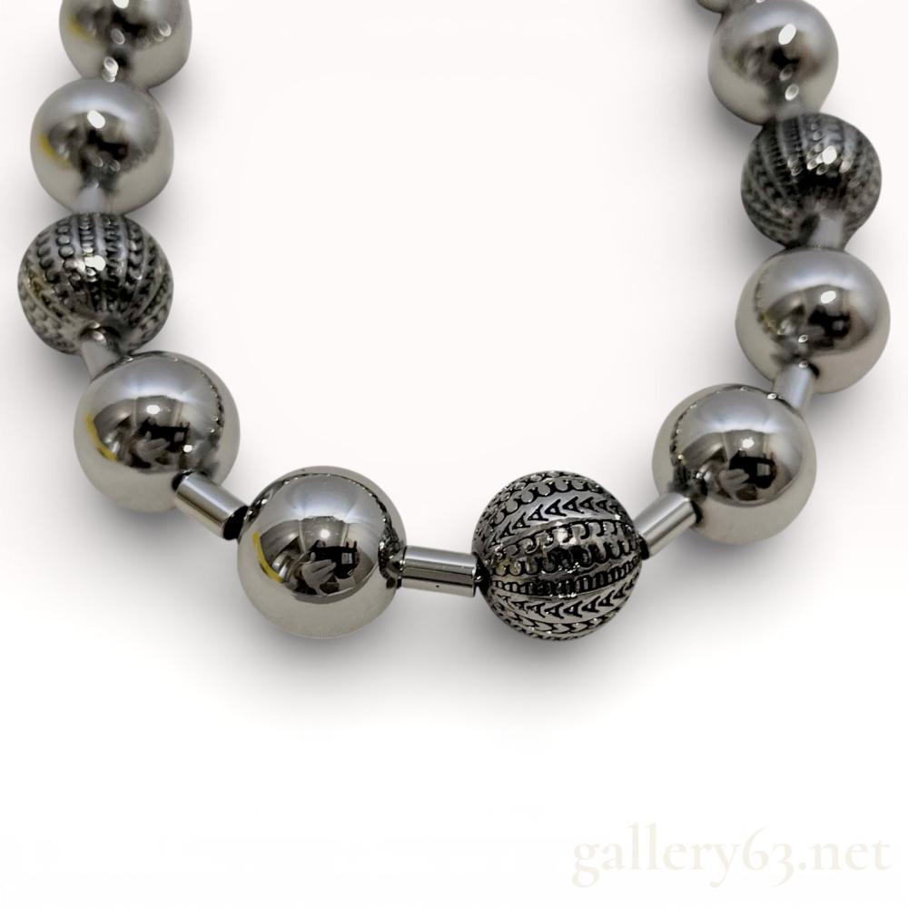 Description
Marc Jacobs necklace composed of a silver-tone ball chain featuring alternating smooth and textured spheres. Select beads engraved with repeating monogram-style patterning creating visual contrast. Polished metal finish throughout. Necklace forming a bold, rhythmic line around the neck.