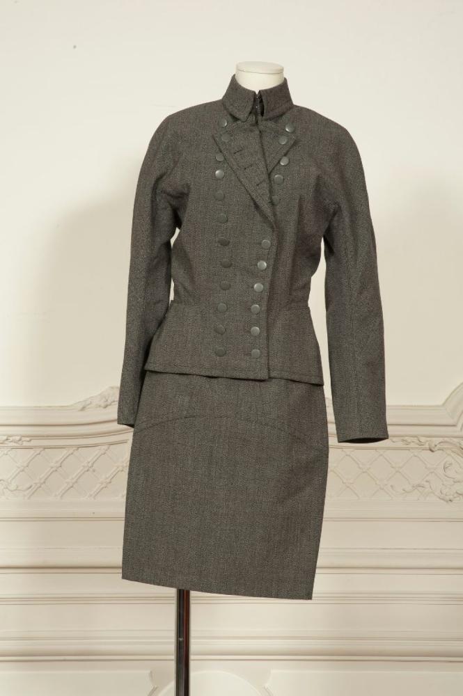ALAIA circa 1990

Tailleur en lainage chiné noir et gris, veste cintré à encolure montante, col rabattu d’inspiration militaire, double boutonnage, poche droite, manches longues, jupe droite à découpe. Griffe blanche, graphisme noir