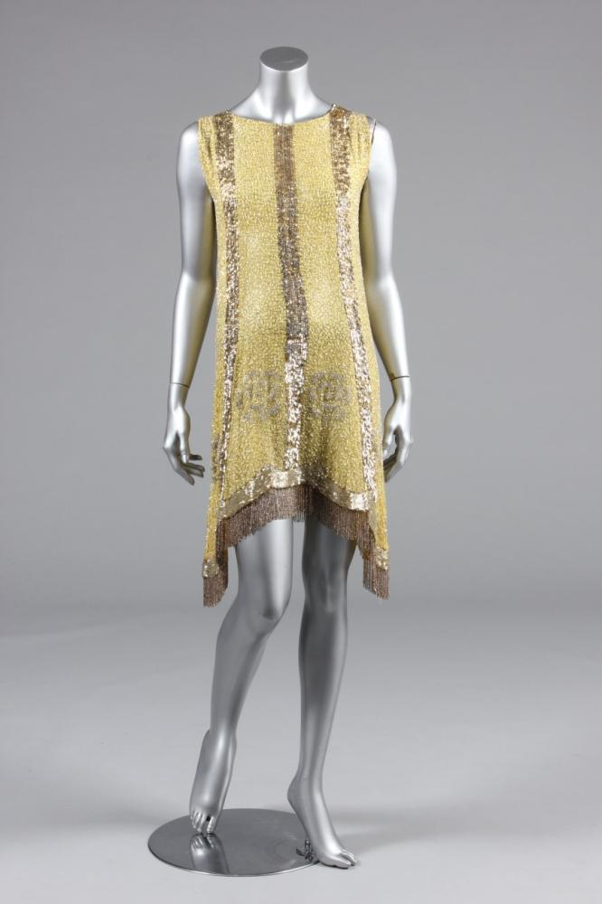 

A beaded primrose-yellow muslin flapper dress, late 1920s, the ground covered with silver vermicular patterning, with seed-bead flowerheads, vertical gold and silver sequined stripes and gold beaded fringes to the hem which dips into points at the sides, bust 86cm, 34in
