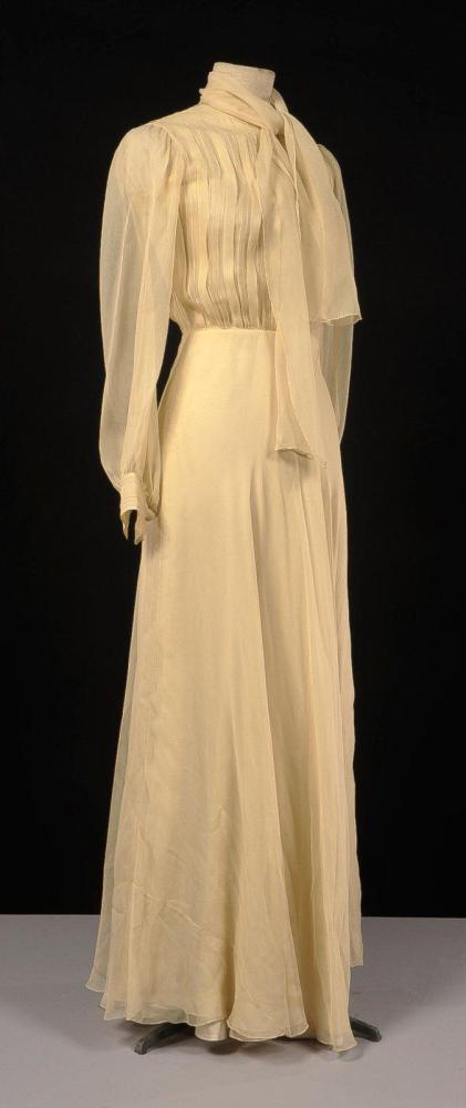 RAFFAELLA, circa 1978

Robe du soir en mousseline de soie beige, haut plissé ras du cou souligné d’un pan faisant cravate, simple boutonnage bride, manches longues ressérés aux poignets, jupe longue travaillée en biais sur double épaisseur sur jupon de soie ivoire. Griffe noire, graphisme blanc.