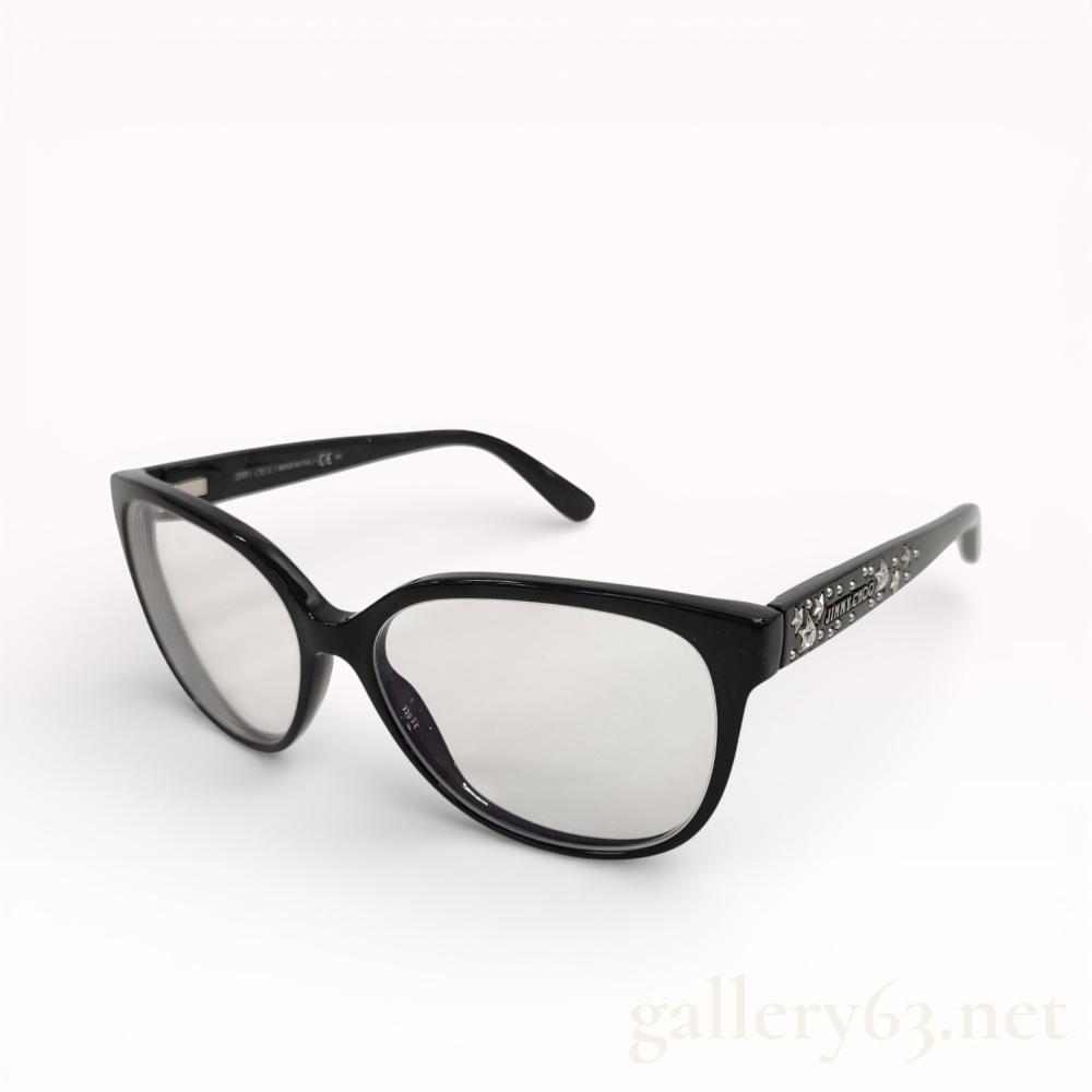 Description
Jimmy Choo model 101 eyeglasses, contemporary. Polished black acetate frames in a modified cat-eye shape featuring silver-tone celestial temple accents with inset crystals and circular studs. Inner temples are marked JIMMY CHOO MADE IN ITALY and JIMMY CHOO 101 29A 135 with raised branding on the exterior. Includes a cream snakeskin-textured cylindrical hard case with silver-tone trim and embossed logo.