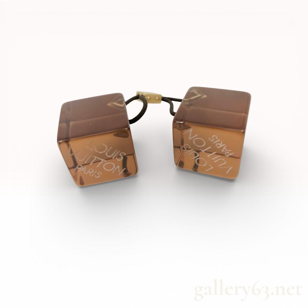 Description
Louis Vuitton hair cubes, contemporary designer accessory. Pair of translucent brown resin cubes featuring etched brand name and LV monogram, connected by a black elastic cord with a gold-tone metal slider. One face of each cube is etched with Louis Vuitton Paris, while another face displays the signature intertwined LV logo.
Each cube: 1 x 1 x 1 inches
Overall length: 5.5 inches.