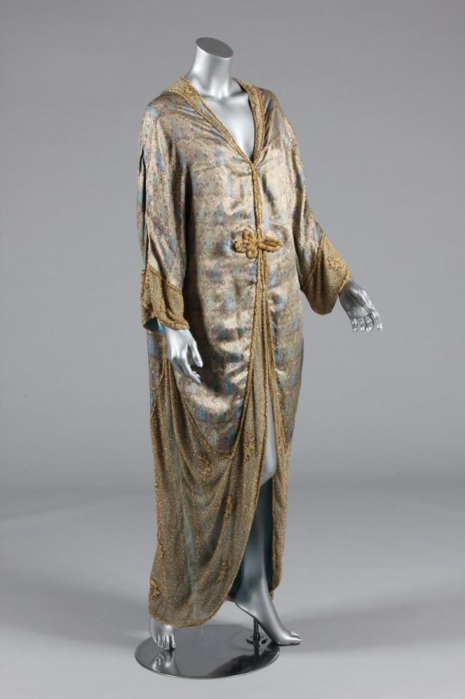 

A Peter Robinson of Regent St, brocaded and beaded cocoon shaped opera coat, early 1920s, with remains of woven label to the blue silk lining, of Persian style brocade with gold and clear glass beaded collar, fastener and hem, beaded fringe to collar edge
