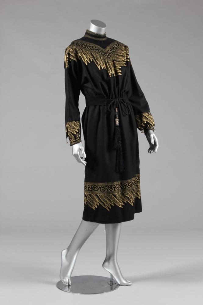 

A Zandra Rhodes black ultra-suede and wool cocktail dress, circa 1980, labelled, boldly printed in gold, the cuffs trimmed with gilt bells, with matching belt, bust approx 86-92cm; together with a gold embroidered black Palestinian jacket c.1950 (3)
