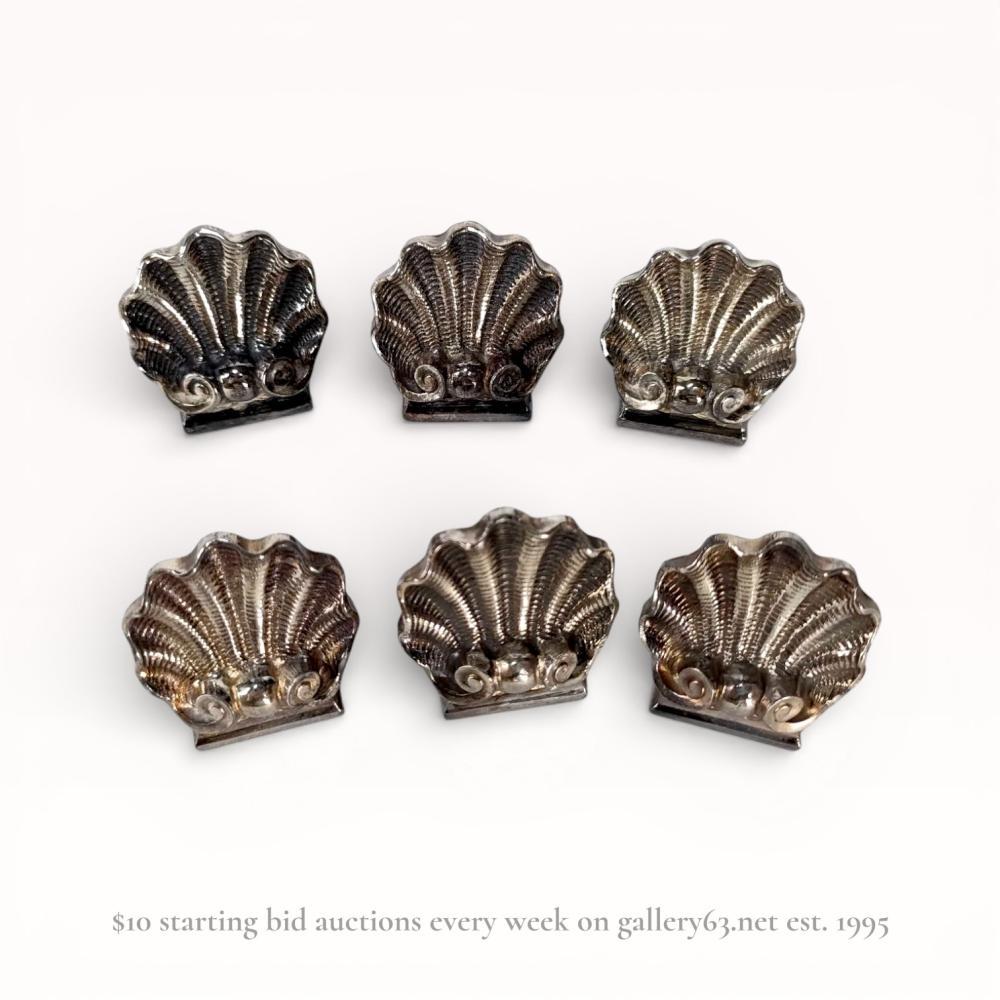 Description
A set of six silver-plated place card holders by Christofle, France, 20th century. Each holder is modeled as a ribbed scallop shell with a scrolled base and a weighted flat bottom designed to secure card stock. The undersides are stamped CHRISTOFLE and FRANCE in block letters with a square maker mark featuring a balance scale flanked by O and C under a star. Includes the original green presentation box marked Christofle Orfevre a Paris.
Width: Approximately 3 cm
Height: Approximately 2.5 cm.