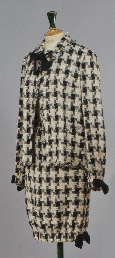 CHANEL, circa 2004

Ensemble en tweed coton polyester beige noir à effet de carreaux, veste à petit col noué par des liens de jersey noir, parmentures bord à bord, manches longues à poignets resserrés; jupe droite entravée également rehaussé d’un noeud