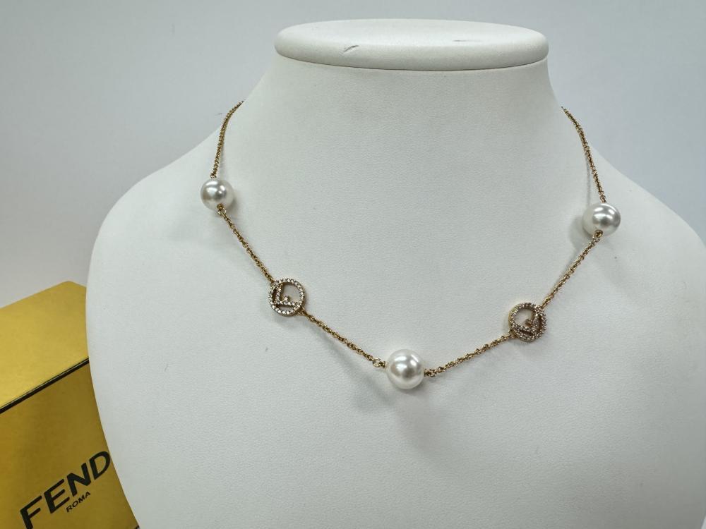 Description
Fendi gold-tone station necklace from the F Is Fendi collection. The fine cable chain is punctuated by four round white simulated pearls and two circular charms featuring the signature F logo bordered by pave-set clear crystals. This item includes an adjustable extender chain with a lobster claw clasp and a terminal logo charm, accompanied by a signature yellow box marked FENDI ROMA.