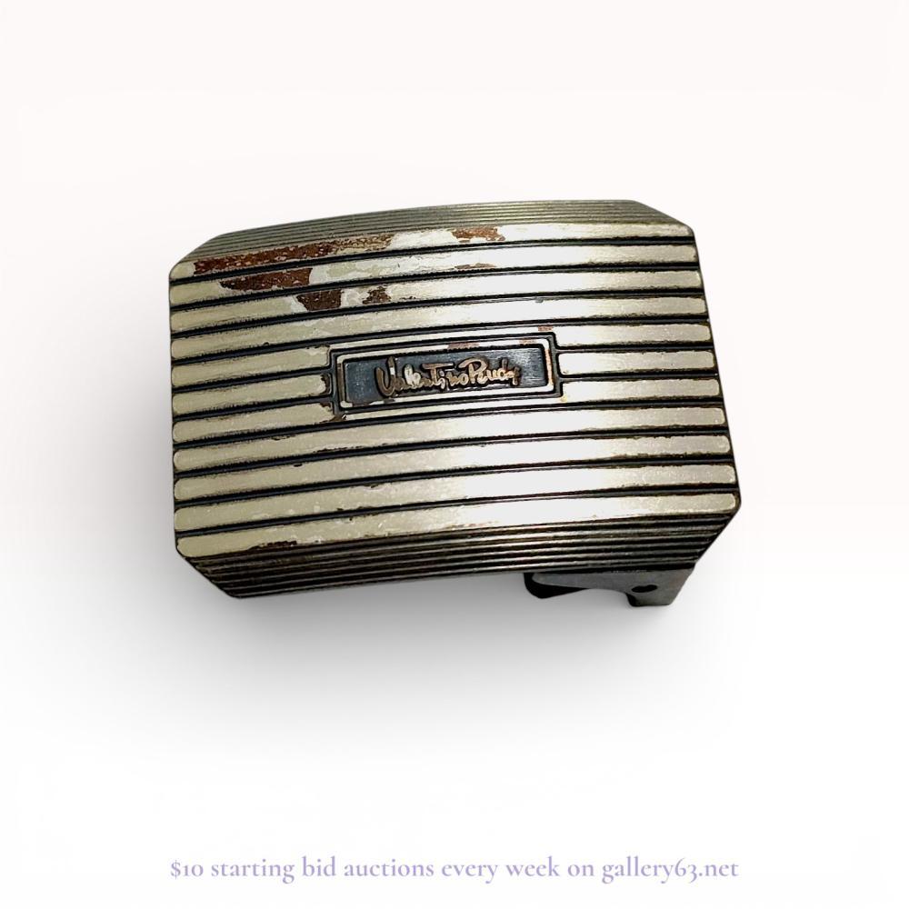 Description
Valentino Rudy plaque-style belt buckle featuring a horizontal ribbed design. The rectangular face includes a central recessed plaque with the brand name in script lettering. Construction includes a fixed bar and a ball-topped peg on the reverse, which is stamped silver.
Approximately 5 cm x 3.3 cm.