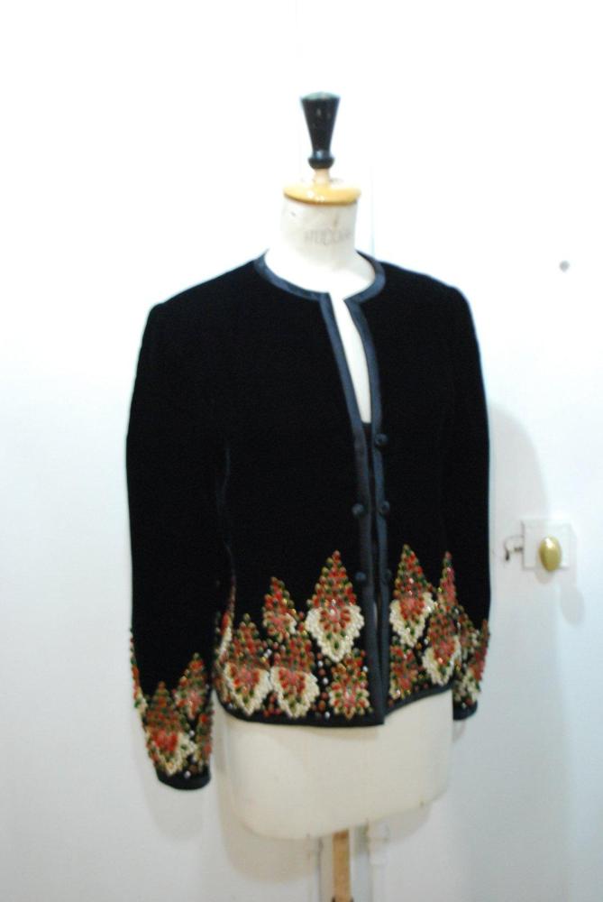 VALENTINO Couture, circa 1985

Veste du soir courte en velours de soie noir gansé de satin, encolure ronde, effet de petites modestie à double boutonnage, bas de la veste et poignets des manches longues rebrodés de strass à l’imitation rubis, émeraude, de perles blanches et de fils or s’inspirant de bijoux de la haute joaillerie. Griffe blanche, graphisme marron