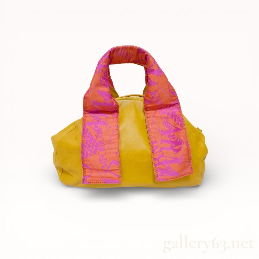 Description
Emporio Armani small satchel bag featuring a smooth yellow leather body and fabric-wrapped top handles. The handles are printed with a repeating pink and orange logo motif, and the interior is lined in a pink and grey striped fabric with eagle logos and GA initials. Construction includes a yellow plastic top zipper with a silver-tone metal eagle logo pull and one interior zippered pocket.
Approximately 11.5 inches width at base.