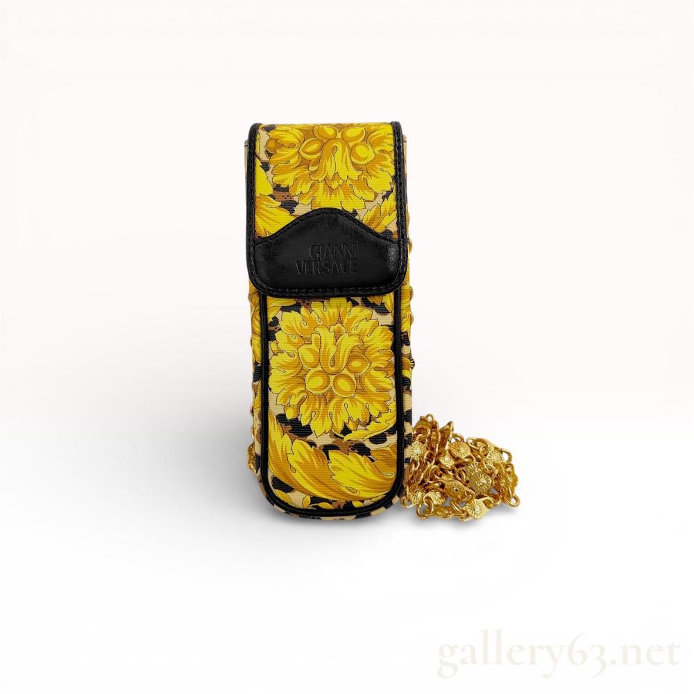 Description
A vintage Gianni Versace mini shoulder pouch, designed as a mobile phone case or accessory pochette. This 1990s piece features a dual-patterned design with a vibrant yellow baroque floral motif on the front and a classic leopard print on the reverse and sides. The exterior is crafted from printed PVC with black leather trim. It features a long, ornate gold-tone chain strap with circular links embossed with a sunburst motif. The pouch is secured with a black leather flap featuring a Velcro closure and a gold-foiled brand signature on the interior. Includes the original Gianni Versace branded box. Dimensions: Approximately 18cm H x 7.5cm W.