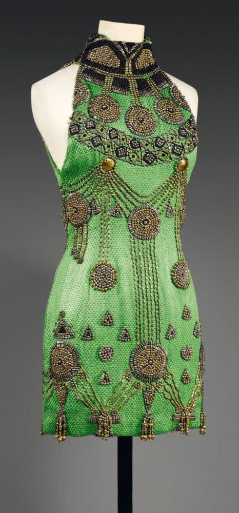 Atelier VERSACE, circa 1985

Mini robe de cocktail en soie tissée vert rebrodée deperlesrondes, tubulaires en métal doré vieilli de petites perles de verre noire, encolure montante formant collerette à motisf ethniques, emmanchures américaines, jupe droite. (manques) Griffe noire, graphisme blanc voir reproduction Pl. 42