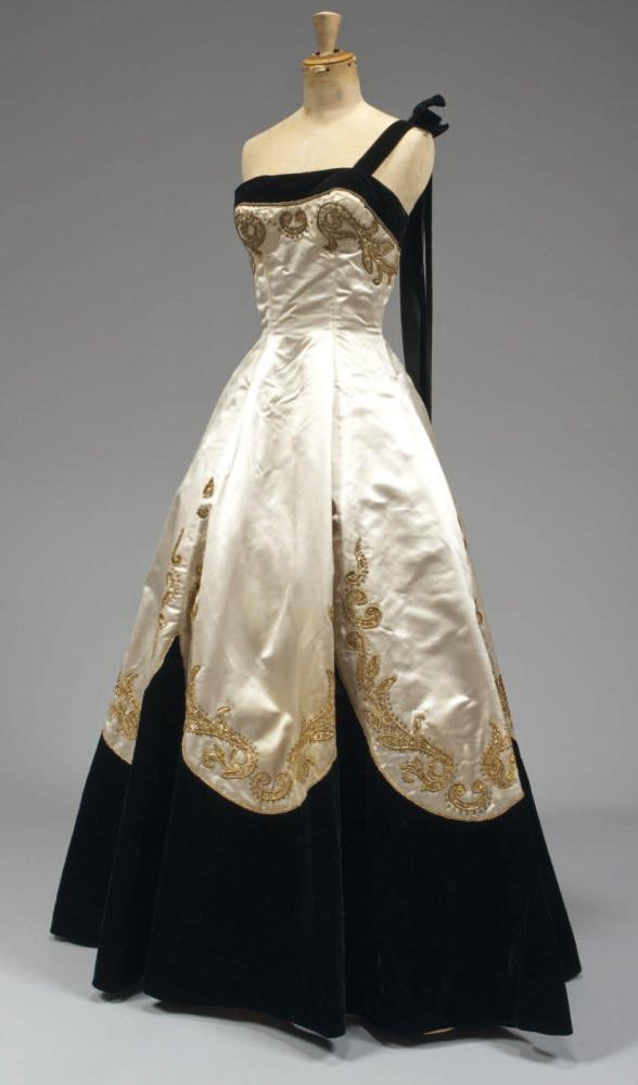 Emilio SCHUBERT Roma, circa 1950

Robe grand soir , haut bustier en satin ivoire brodé de fils or et pailletes soulignant la poitrine rehaussée de velours noir, une bretelle à l’identique; jupe très ample à effet de palmes sur velours avec rappel de la broderie .(bretelle et noeud non d’origine, percale intérieur en l’état) Griffe blanche, graphisme noir