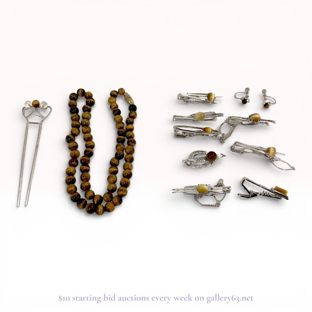 Description
Mid-century collection of tiger’s eye jewelry and accessories including a necklace, hair pin, brooch, earrings, and tie clips. The items feature polished tiger’s eye beads and cabochons displaying characteristic chatoyancy set in silver-tone and gold-tone metal. The kanzashi-style hair pin is stamped SILVER on the reverse, while the necklace features a gold-tone barrel clasp and the earrings utilize screw-back mechanisms.