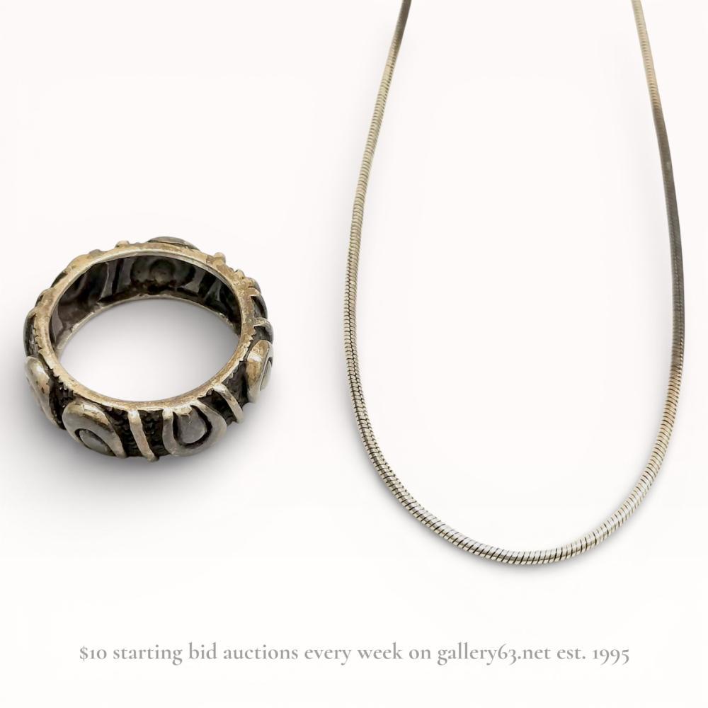 Description
A two-piece sterling silver jewelry group featuring a snake chain necklace and a wide artisan band ring. The necklace is equipped with a spring ring clasp and curb chain extender, with 925 stamps located on the end connector and clasp. The ring displays raised abstract scroll motifs against a stippled, oxidized background and contains a 925 hallmark within an oval recessed stamp on the interior.
Necklace: 15.25 inches maximum length
Ring: US size 10.25, 8 mm width.