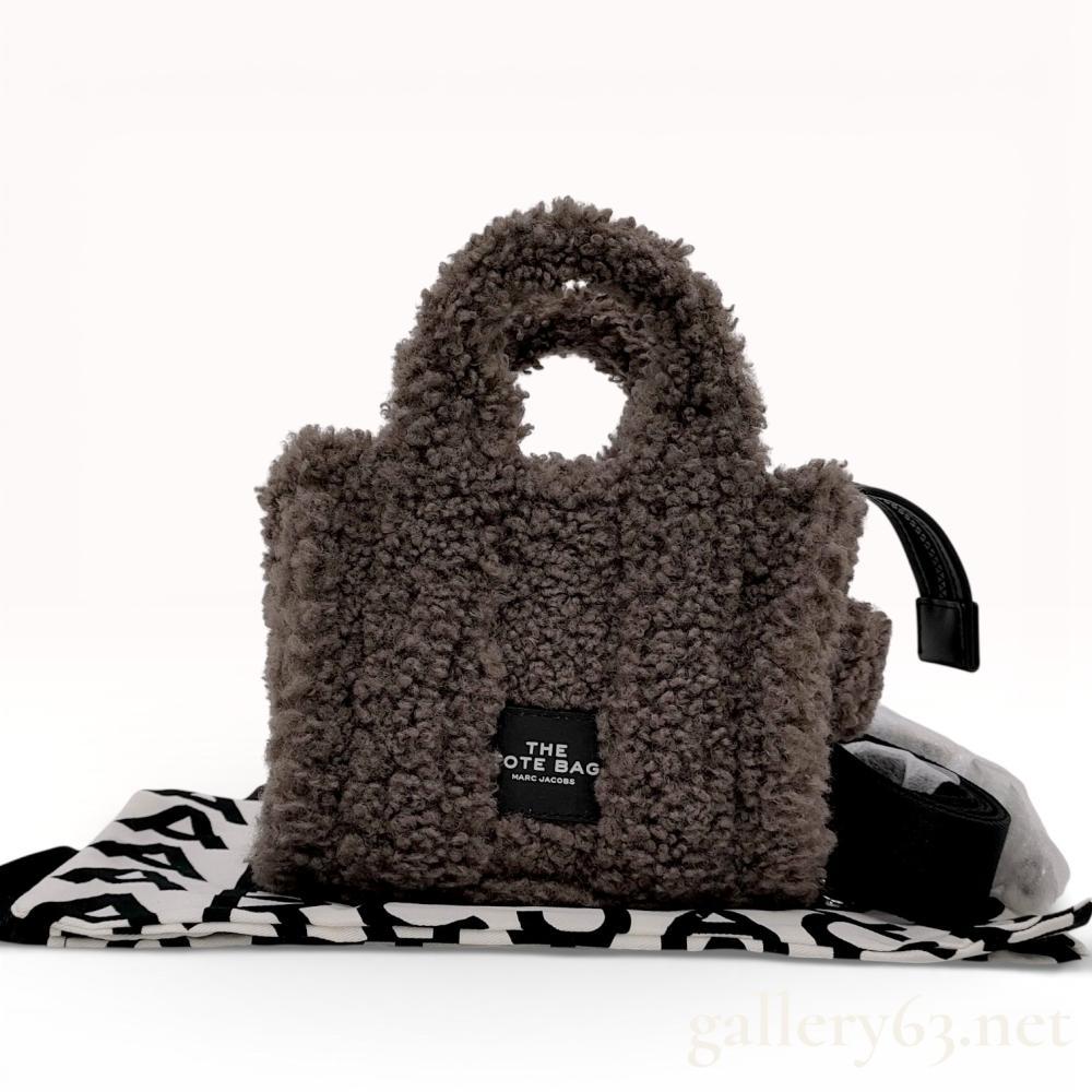 Description
Marc Jacobs The Teddy mini tote bag in charcoal gray faux-shearling fabric. The exterior features dual rolled top handles, a top zip closure with a leather-style pull, and a branded front patch. The interior is lined with gray ribbed textile and includes a black leather-style designer label and fabric care tags.
Base width: approximately 7 inches.