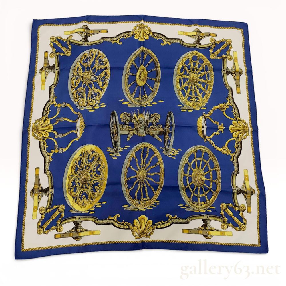 Description
Silk scarf by Hermès, having a square format with hand-rolled edges. Surface featuring a symmetrical carriage wheel and equestrian hardware motif arranged within an ornate border. Palette dominated by deep blue with gold, cream, and black detailing. Design incorporating repeated wheel medallions and classical Hermès-inspired elements. Vintage Hermès "Roues de Canon" silk scarf designed by Caty Latham in 1967.