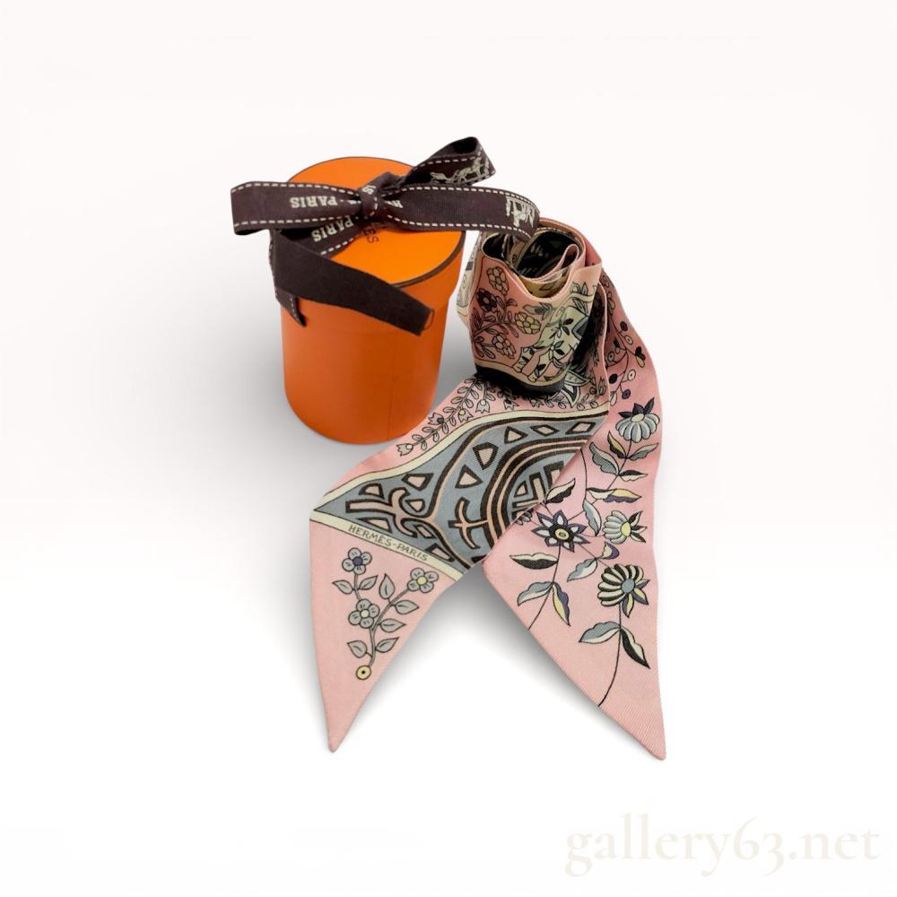 Description
Hermès silk twilly scarf. Narrow format. Pink ground with mixed floral and geometric patterns in black, green, and cream. Printed design alternating along the length. Tapered ends. Hermès Paris marking present. Light wear consistent with handling.