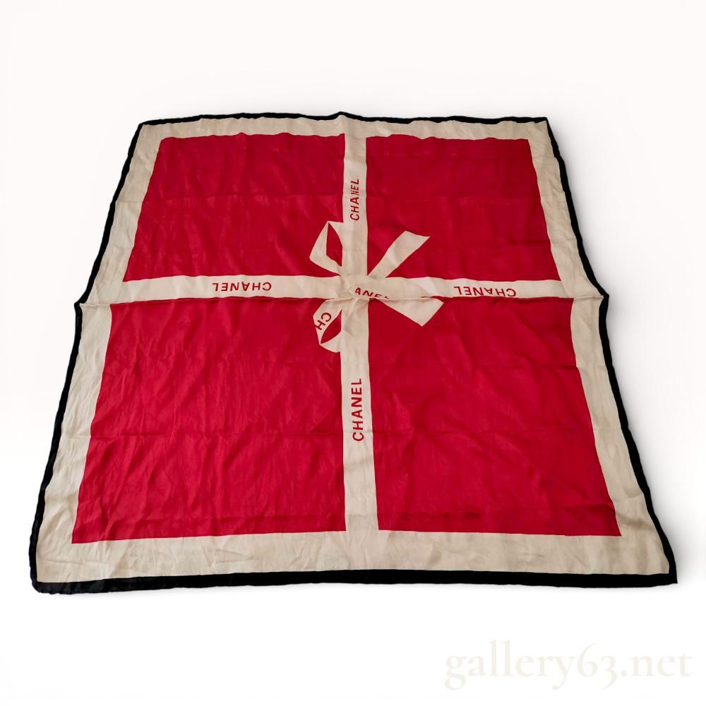 Description
Chanel square silk scarf featuring a gift box motif. The design consists of a vibrant red ground with cream-colored intersecting ribbon bands forming a central bow, finished with a black hand-rolled edge. The Chanel brand name is printed in a red serif typeface repeatedly along the ribbon sections.
Approximately 34 x 34 inches.